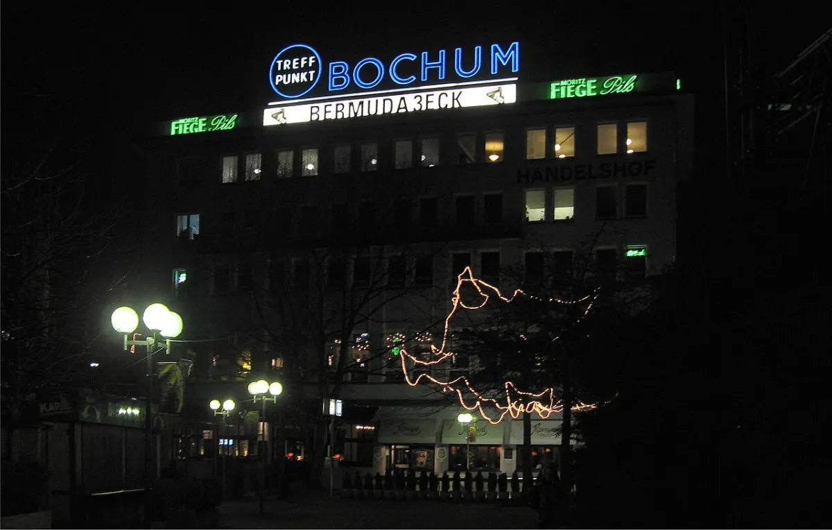 Bei Nacht leuchten auf einem Geschäftshaus blaue und grüne Werbeschriften, davor Kugelleuchten und eine Lichterkette in den Bäumen.