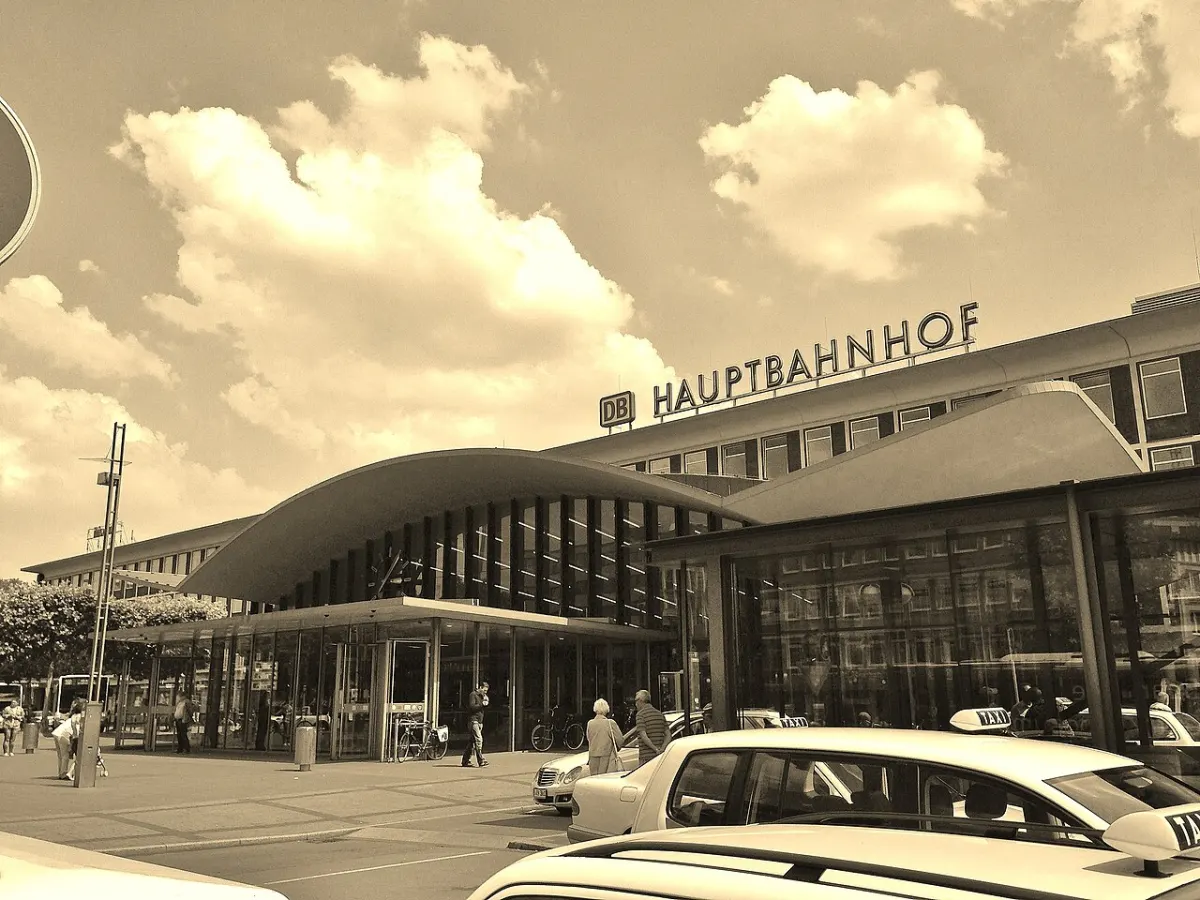 Sepiafarbene Aufnahme eines Bahnhofsgebäudes mit geschwungenem Vordach und großen Glasflächen, davor warten Taxis.