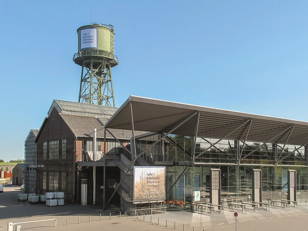 Ein grüner Wasserturm auf Stahlgerüst ragt hinter einer Backsteinhalle mit Glasvorbau und weit auskragendem Stahldach auf.