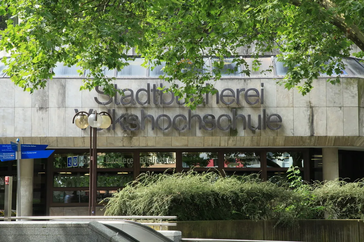 Betonfassade mit großen Metallbuchstaben „Stadtbücherei Volkshochschule“, davor Laub, Sträucher, Laternen und ein blauer Wegweiser.