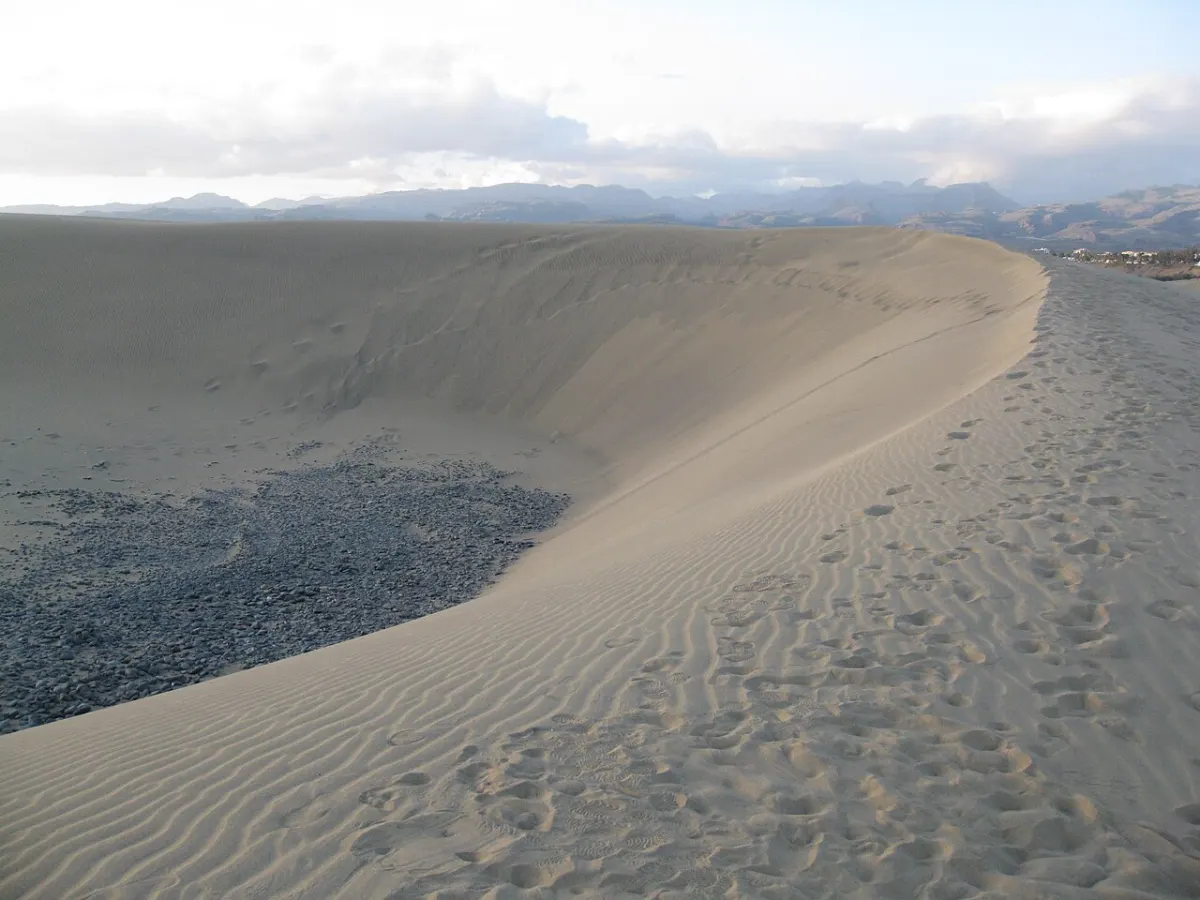 Ein geschwungener Dünenkamm mit gerippeltem Sand und Fußspuren, am Horizont Berge unter Wolken.