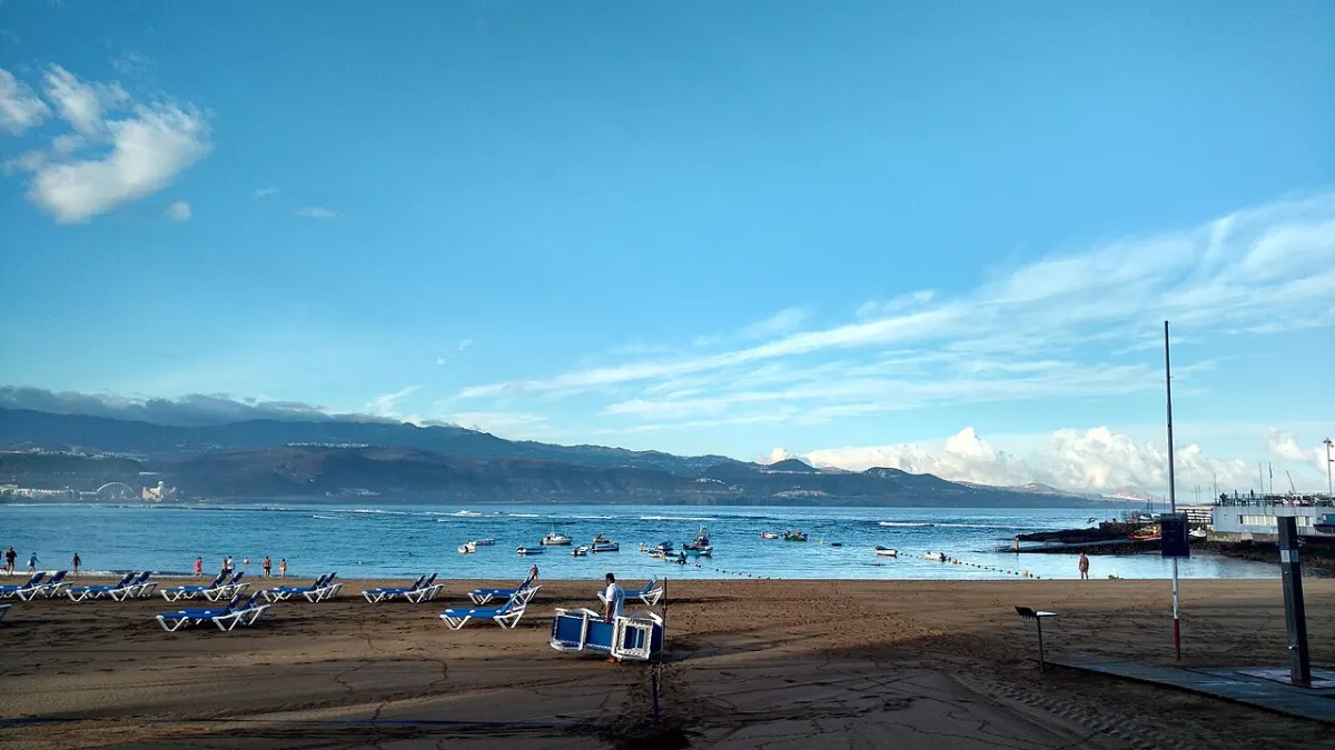 Sandstrand mit Sonnenliegen, davor ruhiges blaues Wasser mit kleinen Booten und am Horizont eine Bergkette unter Schleierwolken.