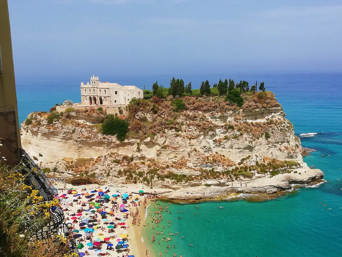 Eine Kirche steht auf einem grün bewachsenen Felsen im türkisblauen Meer, darunter ein Strand voller bunter Sonnenschirme.